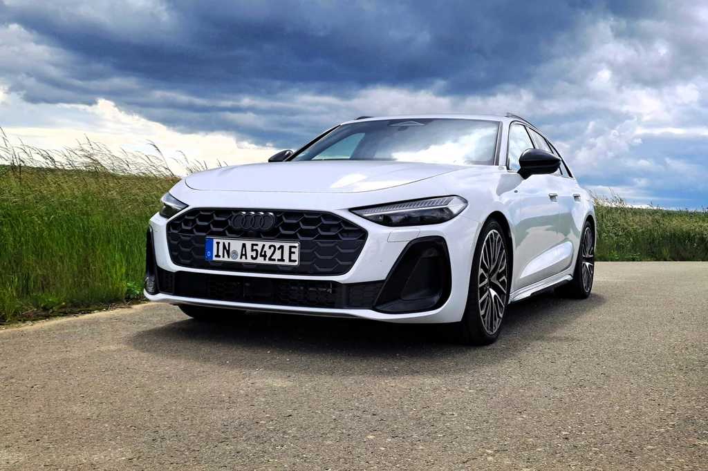 W trybie wyłącznie elektrycznym Audi A5 może rozpędzić się maksymalnie do 140 km/h. Biały samochód marki Audi zaparkowany na asfaltowej drodze, otoczony wysoką trawą, pod zachmurzonym niebem.
