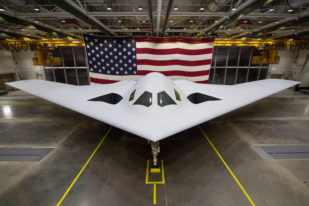 Pocisk LRSO ma być jedną z głównych broni dla najnowszych bombowców stealth B-21 Raider Duży nowoczesny bombowiec strategiczny o futurystycznym kształcie skrzydeł trójkątnych ustawiony jest centralnie w hangarze, przed wielką amerykańską flagą rozwieszoną na tylnej ścianie.