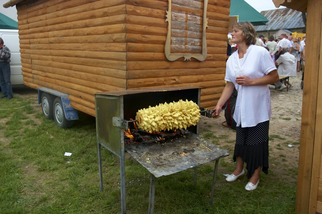 Kobieta stojąca obok tradycyjnego pieca na zewnątrz, obracająca duży sękacz nad otwartym ogniem, w tle drewniany budynek i grupa osób.