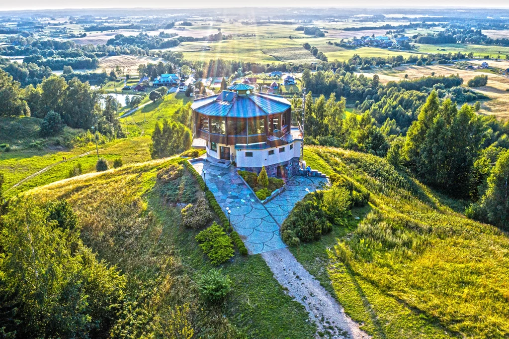 Panorama zielonych wzgórz z dominującym nowoczesnym budynkiem o okrągłym kształcie na szczycie, otoczonym alejkami, a w oddali rozciągają się pola, lasy oraz zabudowania miejscowości.