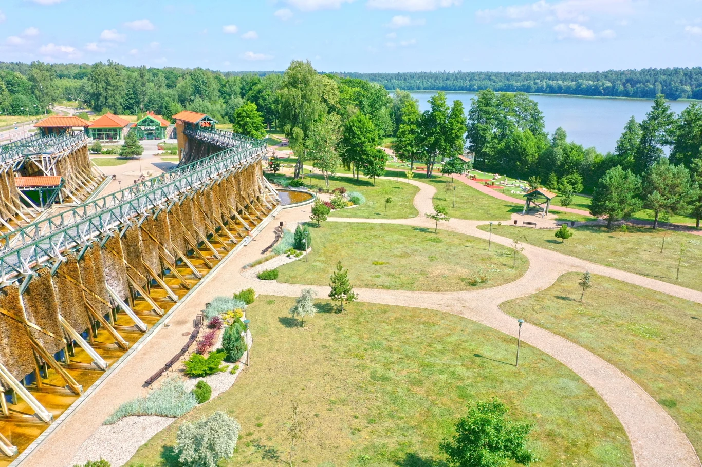 Tężnia solankowa otoczona zadbanym parkiem z alejkami, zielenią i kwiatami, w tle widoczne jezioro i otaczający je las, kilka pawilonów oraz miejsca do wypoczynku.