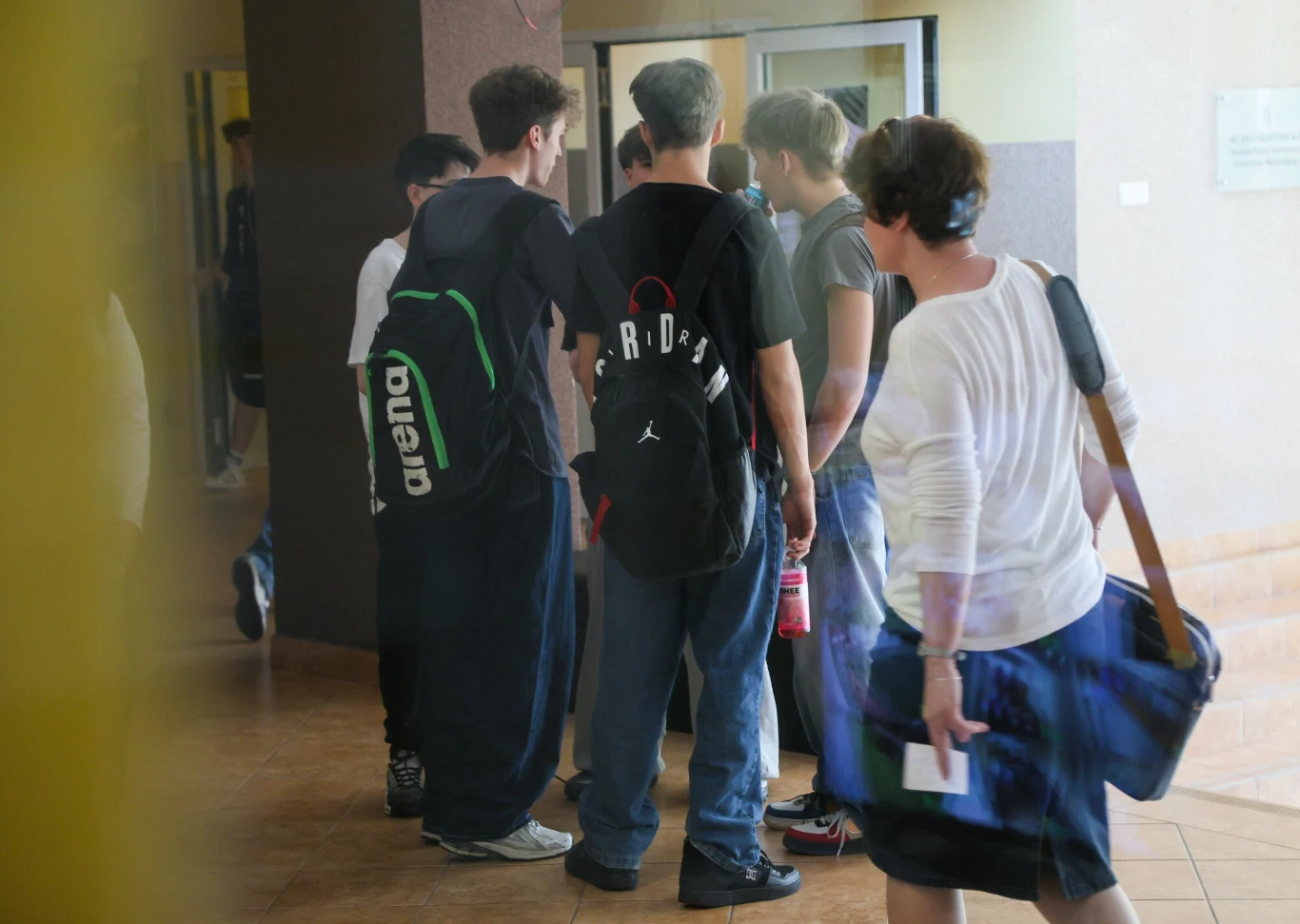 Nauczyciele alarmują: uczniowie mają problem z pisaniem. Sugerują, dlaczego Grupa młodych osób, głównie nastolatków, rozmawia w korytarzu szkolnym, obok przechodzi kobieta z torebką na ramieniu.