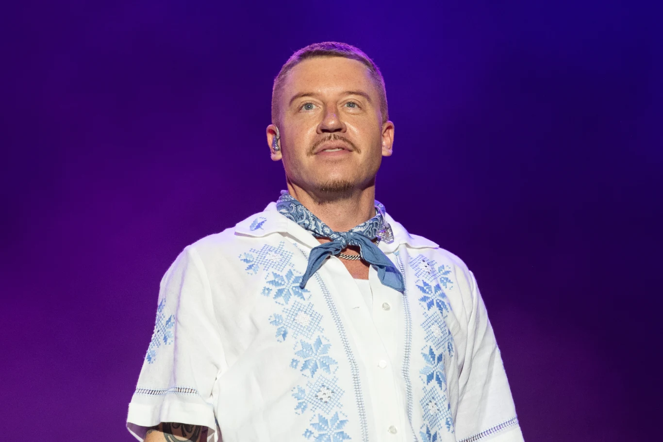 Włamanie w domu Macklemore'a Mężczyzna z krótkimi włosami i wąsem ubrany w białą koszulę z niebieskimi haftami i niebieską apaszką na szyi stoi na tle fioletowego światła.