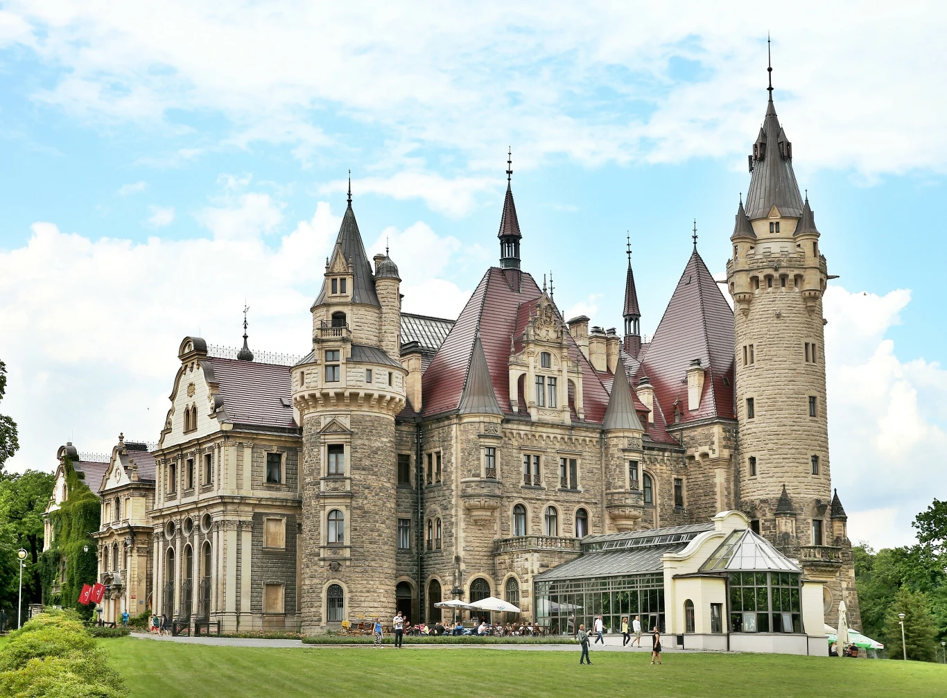 Opolski Hogwart Imponujący zamek o licznych wieżyczkach i detalach architektonicznych, otoczony zadbanym trawnikiem, na którym przebywają ludzie odpoczywający lub spacerujący; po prawej stronie znajduje się przeszklona oranżeria.