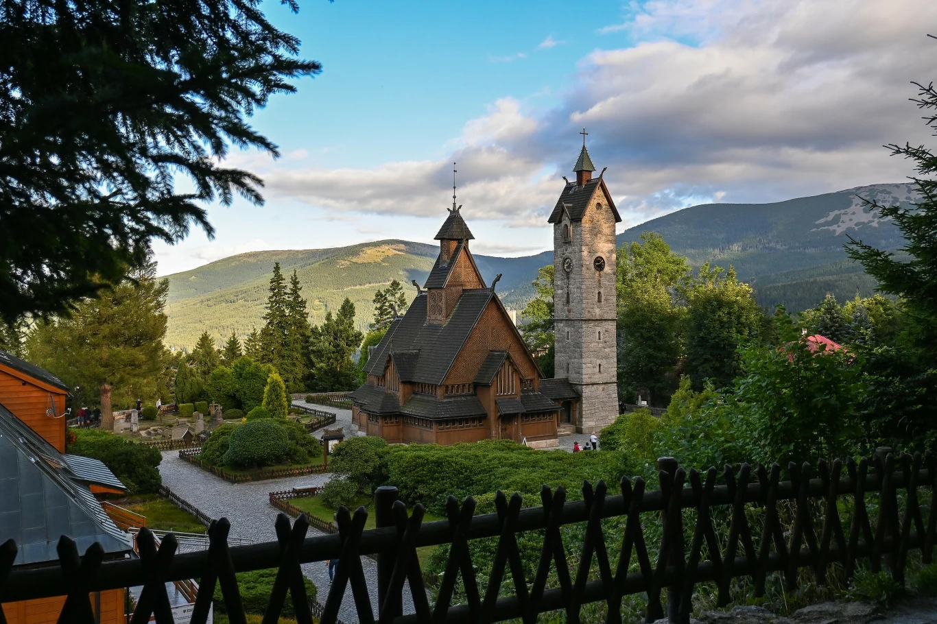 Wyjątkowe kościoły w Polsce Drewniany kościółek z charakterystyczną wieżą oraz osobno stojąca dzwonnica otoczone zielenią, w tle rozciągają się górskie krajobrazy i lasy, na niebie widoczne chmury oświetlone światłem dziennym.