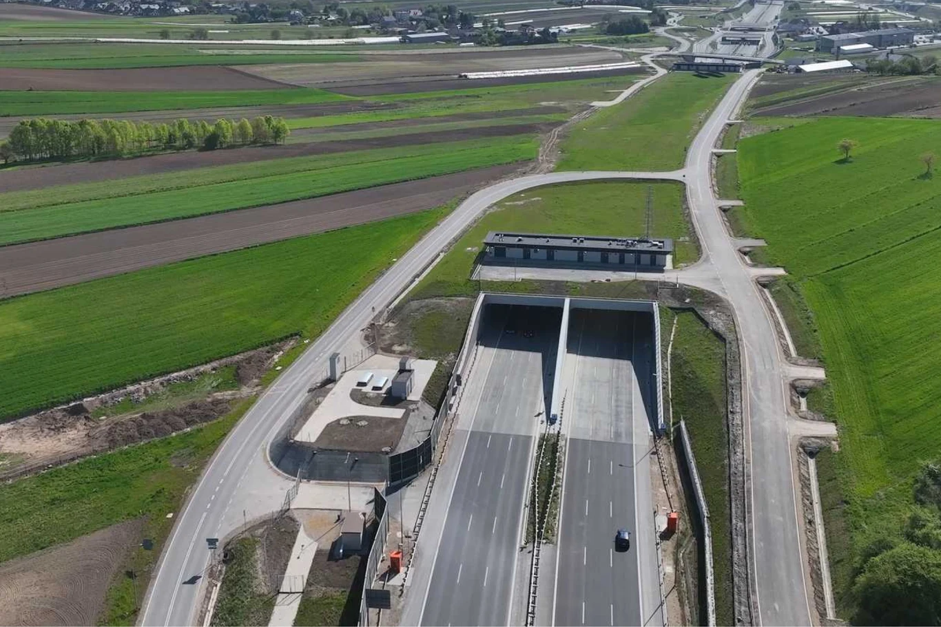 Tunel w Zielonkach mierz 653 m długości. Został otworzony pod koniec ubiegłego roku. Tunel drogowy przebiegający pod wiejskim terenem, dwupasmowa jezdnia z nielicznymi pojazdami, otoczona polami uprawnymi i bocznymi drogami, w tle widoczne zabudowania oraz oddalone skrzyżowanie.