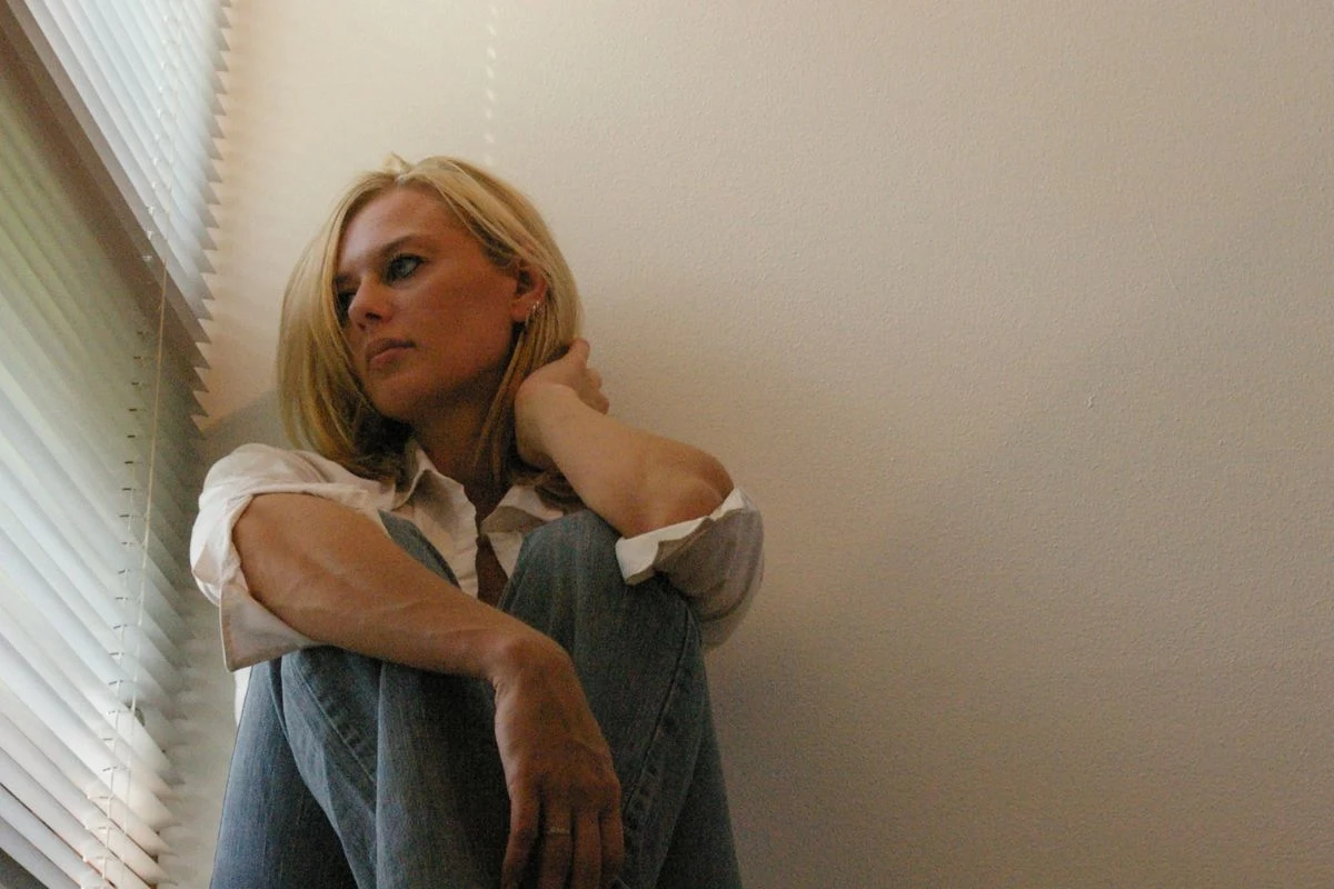 Czy każdy potrzebuje terapii? Kobieta o blond włosach siedzi na podłodze pod białą ścianą, oparta o kolano, patrzy w bok z zamyślonym wyrazem twarzy, ubrana w jasną koszulę i dżinsy, przy oknie z żaluzjami przepuszczającymi światło.