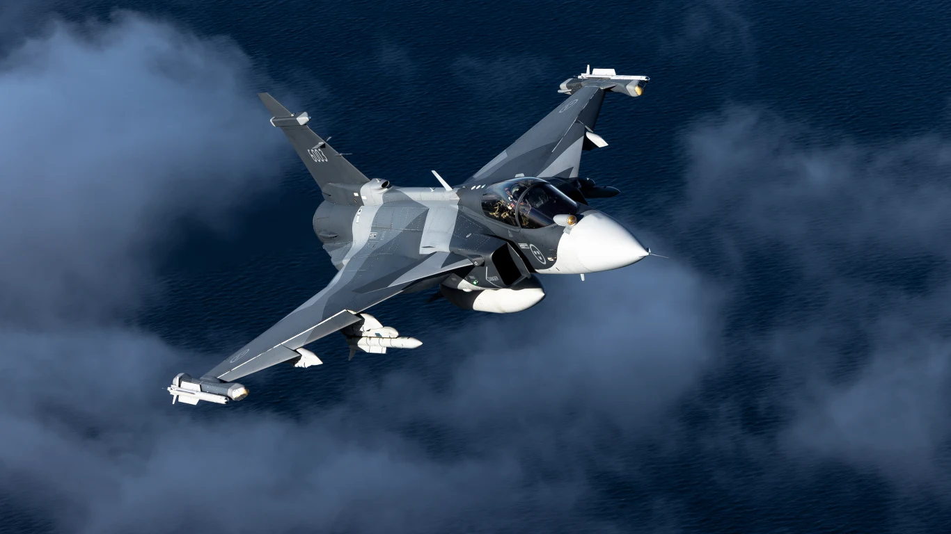 Szwedzi po raz pierwszy przetestowali myśliwiec wspierany sztuczną inteligencją w walce z człowiekiem Wojskowy samolot myśliwski o nowoczesnej sylwetce przelatuje nad gęstymi chmurami, wyraźnie widoczne uzbrojenie pod skrzydłami oraz kokpit z pilotem.