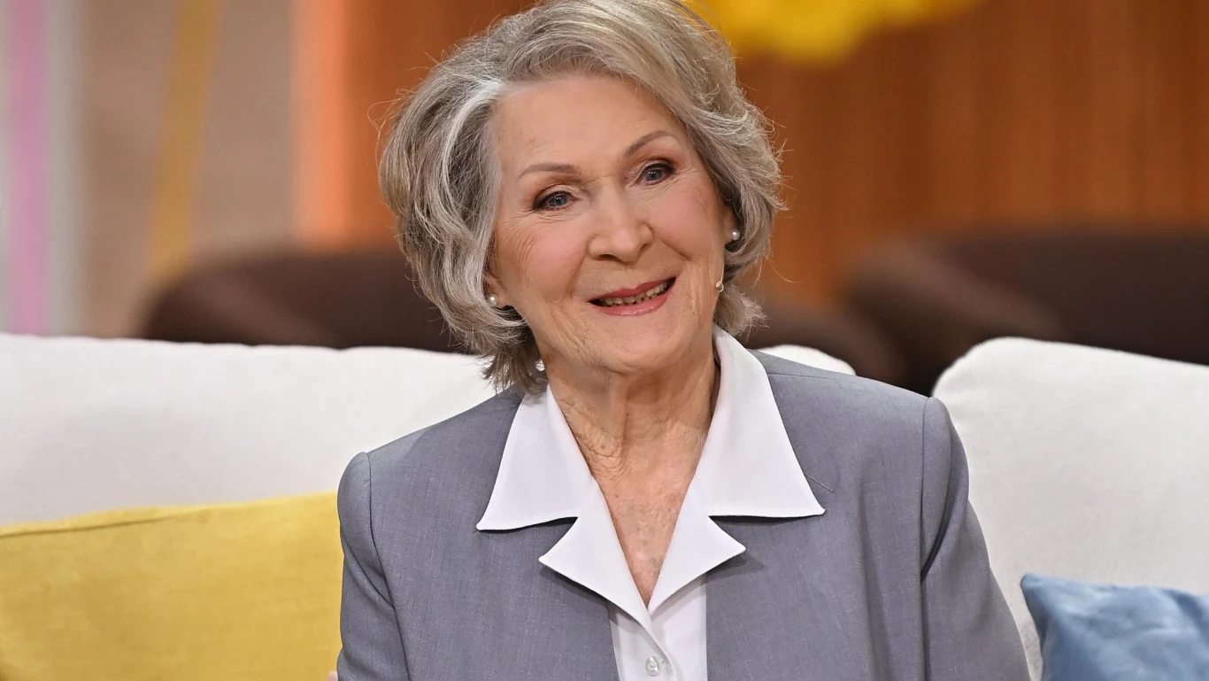 Irena Santor Starsza kobieta z uśmiechem siedzi na jasnej sofie, ubrana w elegancki żakiet i białą koszulę, w tle rozmyte wnętrze z żółtymi i brązowymi akcentami.