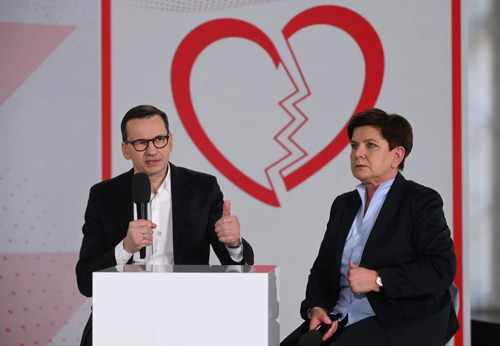 Byli premierzy: Mateusz Morawiecki i Beata Szydło Dwoje dorosłych w formalnych strojach siedzi na tle dużego, czerwonego serca z charakterystycznym pęknięciem pośrodku; mężczyzna trzyma mikrofon i unosi kciuk, kobieta ma poważny wyraz twarzy.