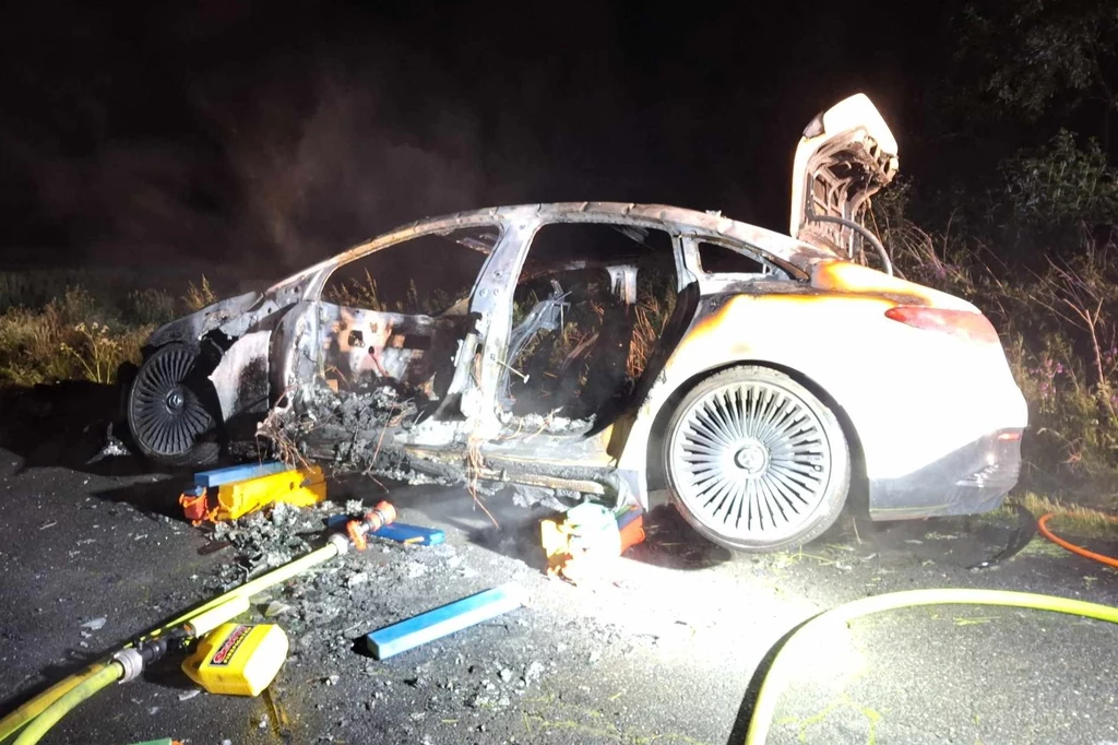 Niewiele zostało ze spalonego Mercedesa EQE. Spalony samochód osobowy stojący na poboczu przy nocnym oświetleniu, pozostałości po pożarze i przetopione elementy, otwarty bagażnik, wokół narzędzia ratownicze i gaśnicze.