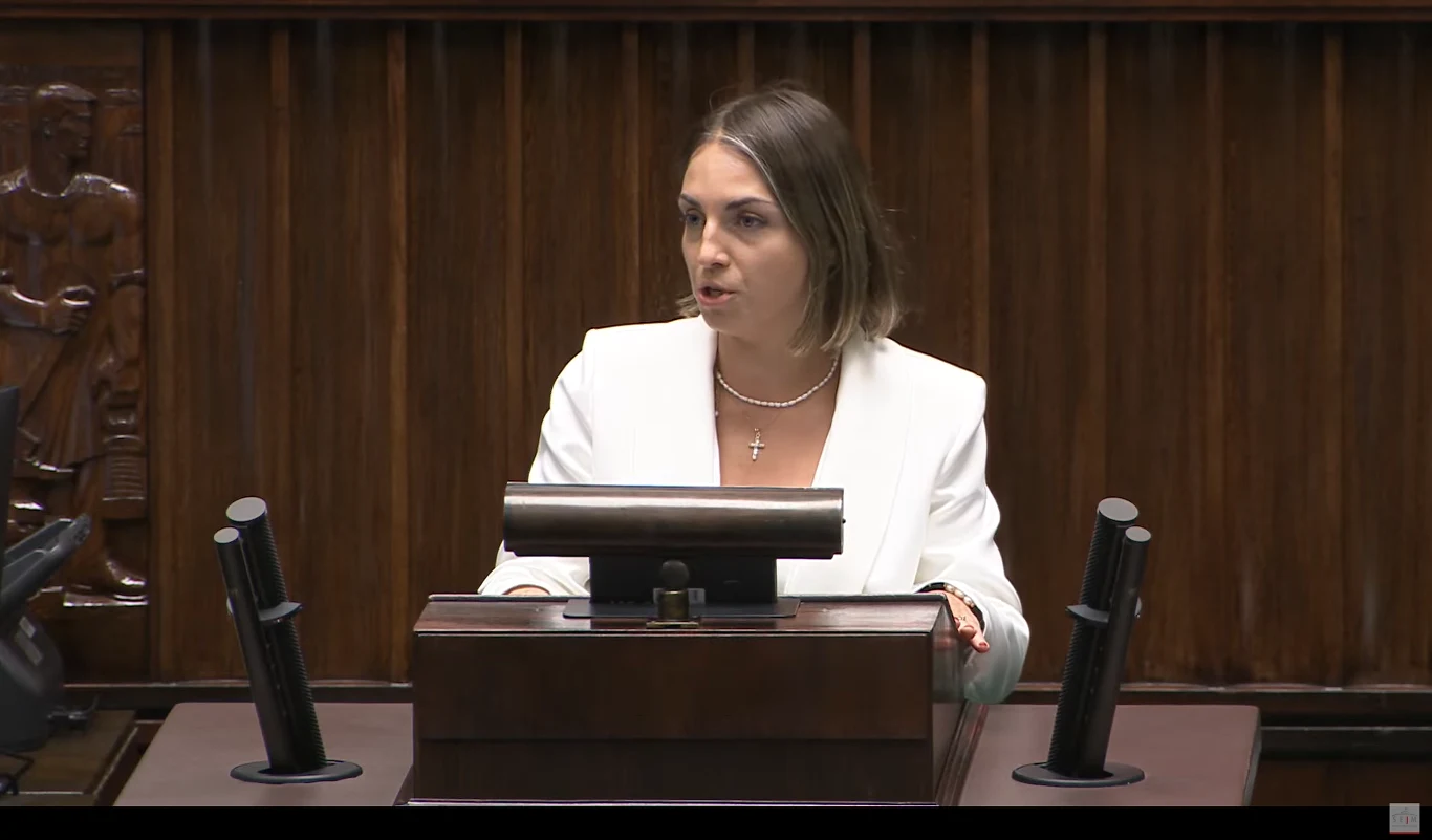 Kinga Gajewska podczas debaty nad wotum zaufania dla rządu Kobieta w białej marynarce stoi przy mównicy w sali o drewnianych panelach, za nią widoczne jest tło z rzeźbionymi detalami, a na mównicy znajdują się mikrofony.