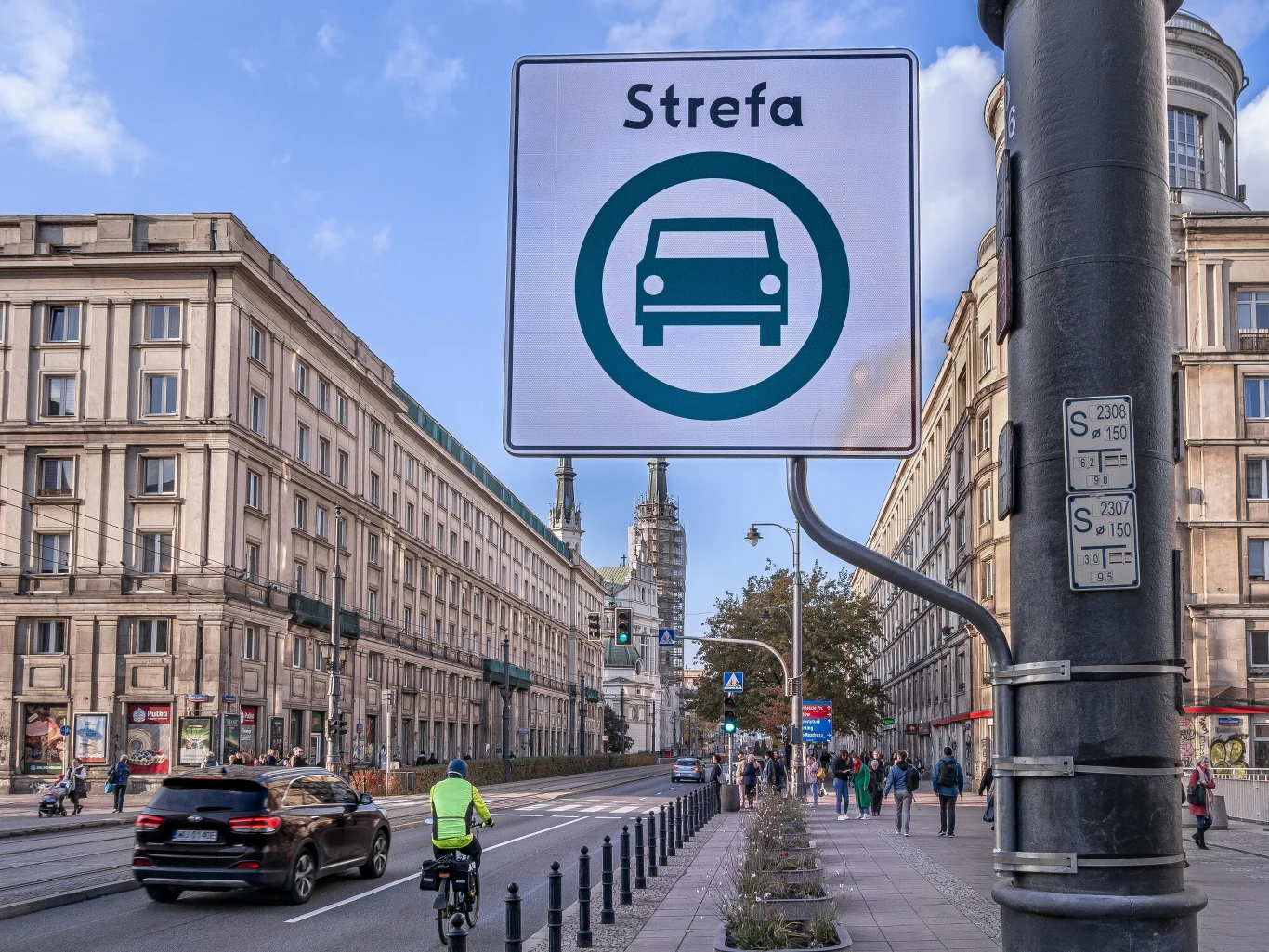 Ulica w centrum miasta z ruchem samochodowym i rowerowym, znak drogowy informujący o specjalnej strefie dla pojazdów, po bokach wysokie kamienice oraz piesi na chodniku, w tle widoczny zabytkowy budynek.