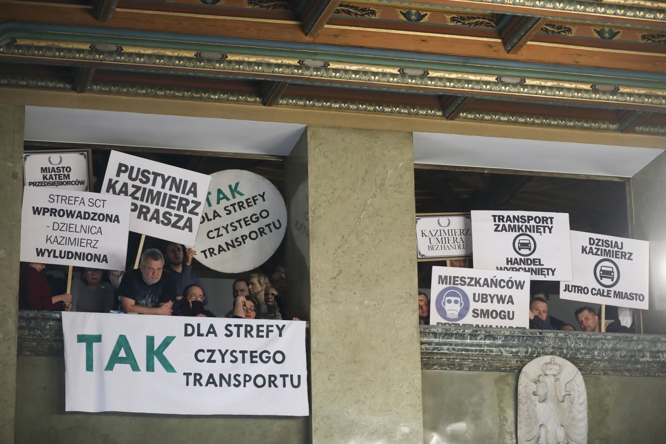 Grupa osób trzyma transparenty i plakaty podczas protestu lub debaty dotyczącej wprowadzenia strefy czystego transportu, z widocznymi hasłami za i przeciw zmianom wpływającym na życie mieszkańców miasta.