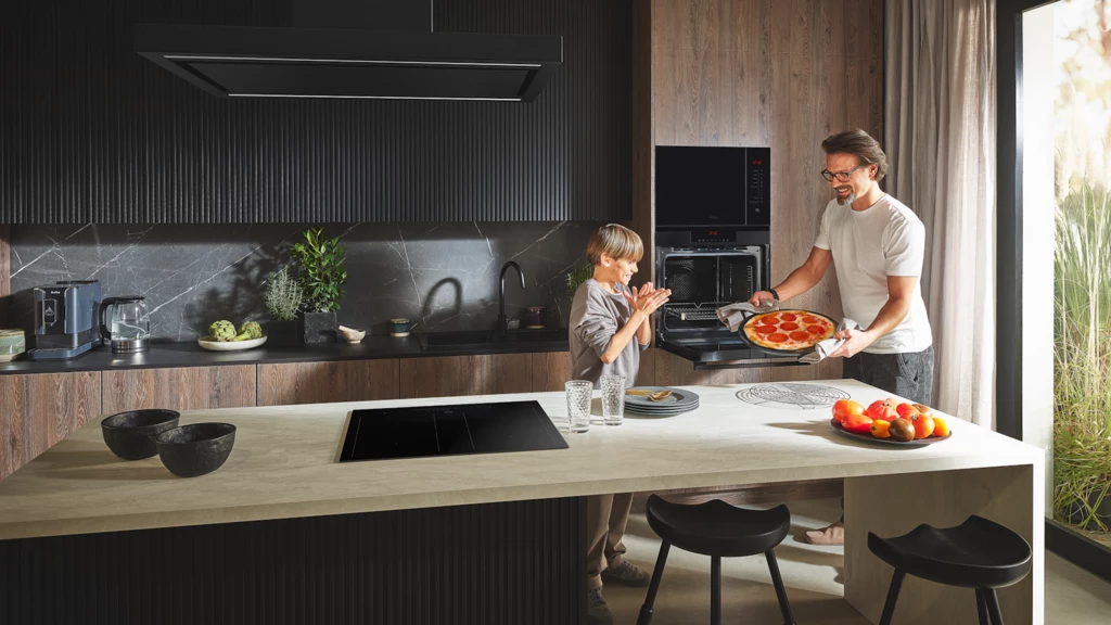 Pieczenie pizzy wymaga cierpliwości, ale odwdzięcza nam się smakiem najpiękniejszych włoskich wakacji Nowoczesna, jasna kuchnia, w której dorosły podaje dziecku pizzę wyjętą z piekarnika, wokół elegancko urządzone wnętrze, na blacie owocowa misa oraz czarne naczynia.