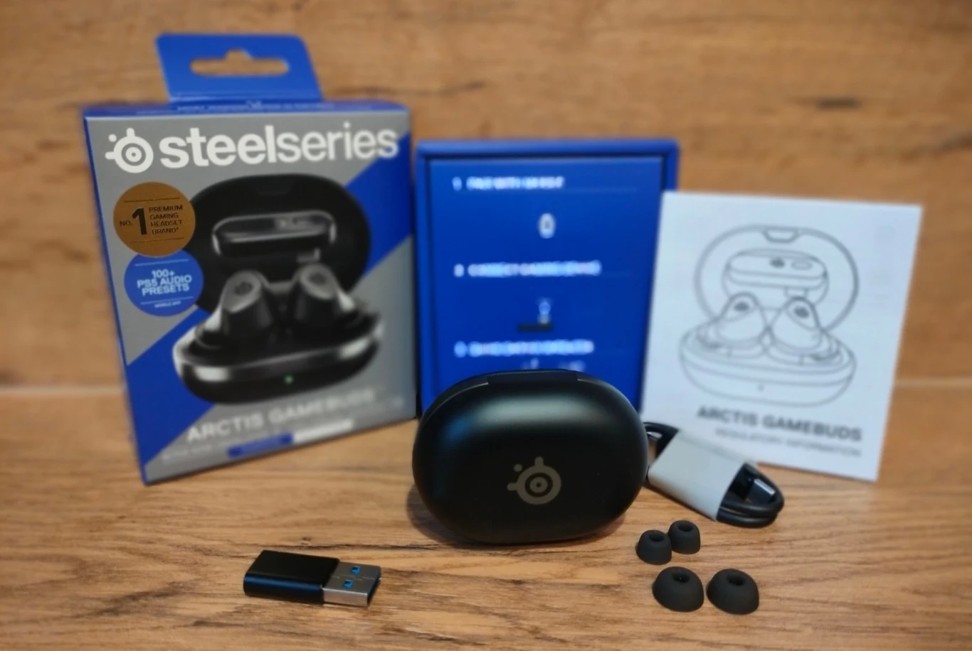Zestaw słuchawek bezprzewodowych SteelSeries Arctis Gamebuds z etui ładującym, opakowaniem, adapterem USB, dodatkowymi wkładkami dousznymi oraz instrukcją obsługi na drewnianym tle.