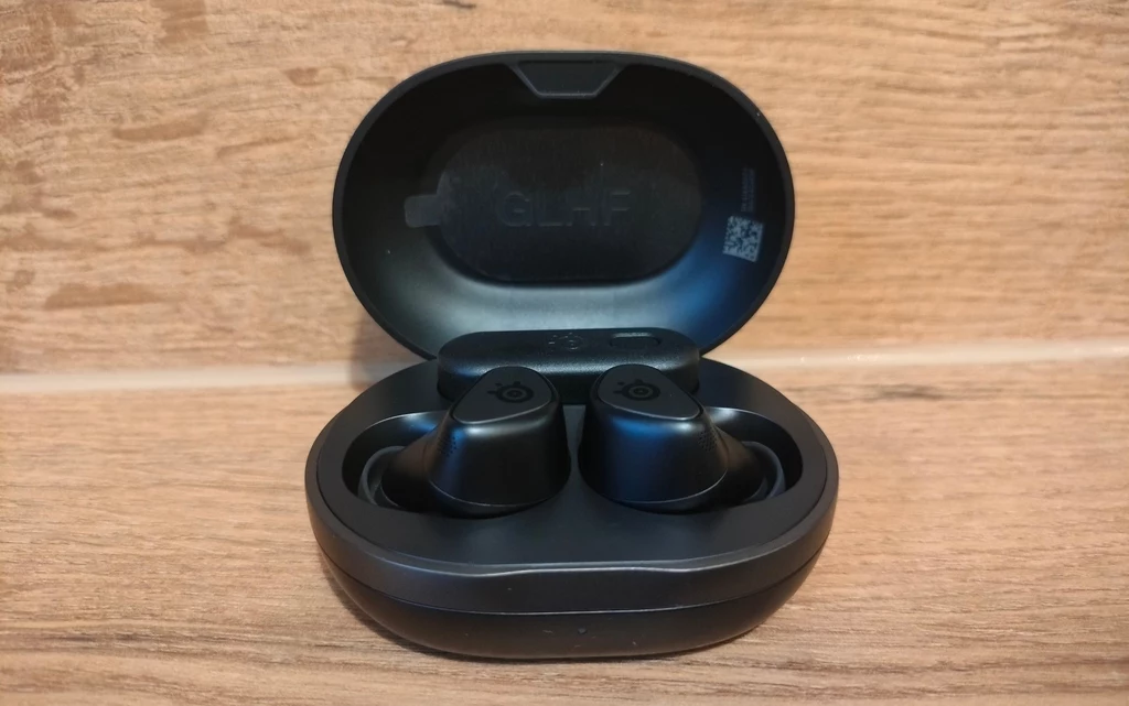 SteelSeries Arctis GameBuds Czarne bezprzewodowe słuchawki douszne umieszczone w otwartym etui ładującym na tle drewnianej powierzchni.