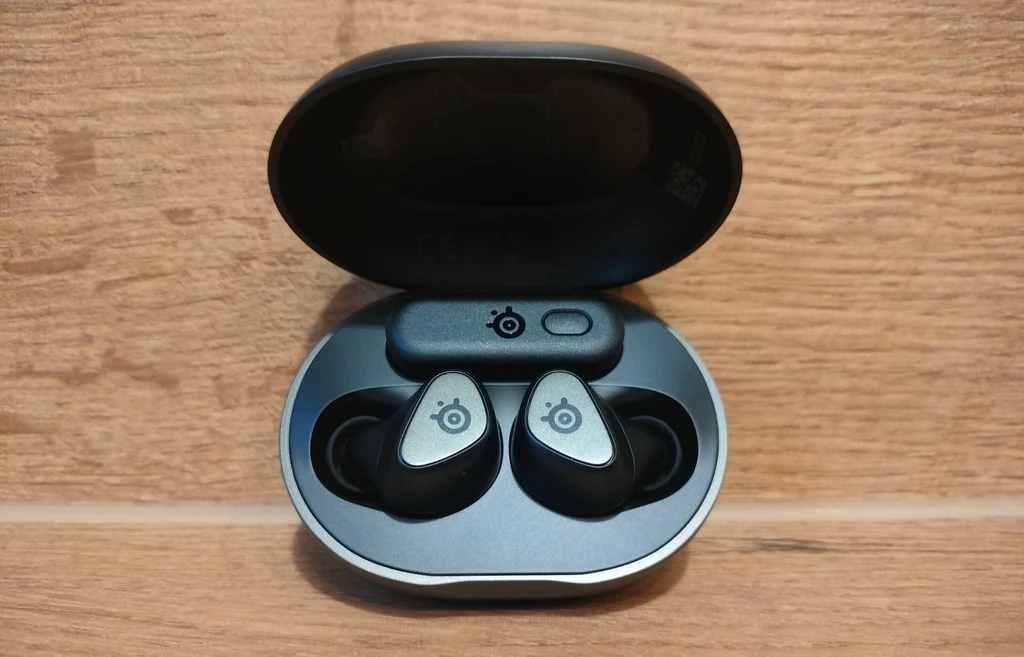 SteelSeries Arctis GameBuds Bezprzewodowe słuchawki dokanałowe umieszczone w otwartym etui do ładowania, ustawione na drewnianej powierzchni, logo producenta widoczne na każdej słuchawce.