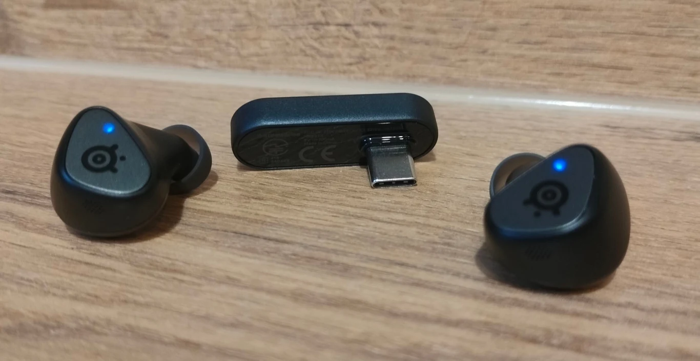Dwie bezprzewodowe słuchawki douszne z niebieskimi diodami LED oraz adapter USB-C umieszczone na drewnianej powierzchni.