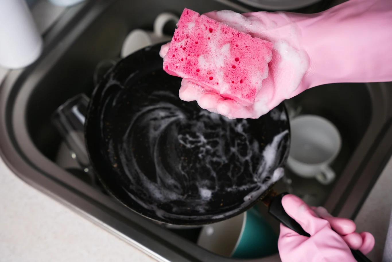 Ręka w różowej gumowej rękawiczce trzyma różową gąbkę pokrytą pianą, myje czarną patelnię w zlewie kuchennym, w tle widoczne są inne naczynia.