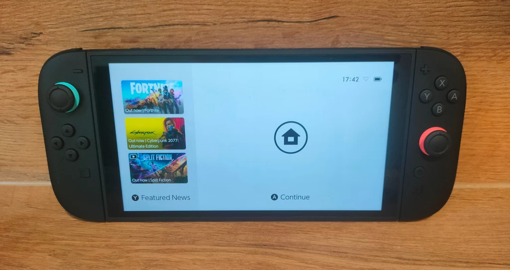 Nintendo Switch 2 Konsola do gier Nintendo Switch leżąca na drewnianej powierzchni, ekran urządzenia wyświetla menu z aktualnościami oraz ikoną przejścia do ekranu głównego, po lewej stronie widoczne kolorowe przyciski joysticka.