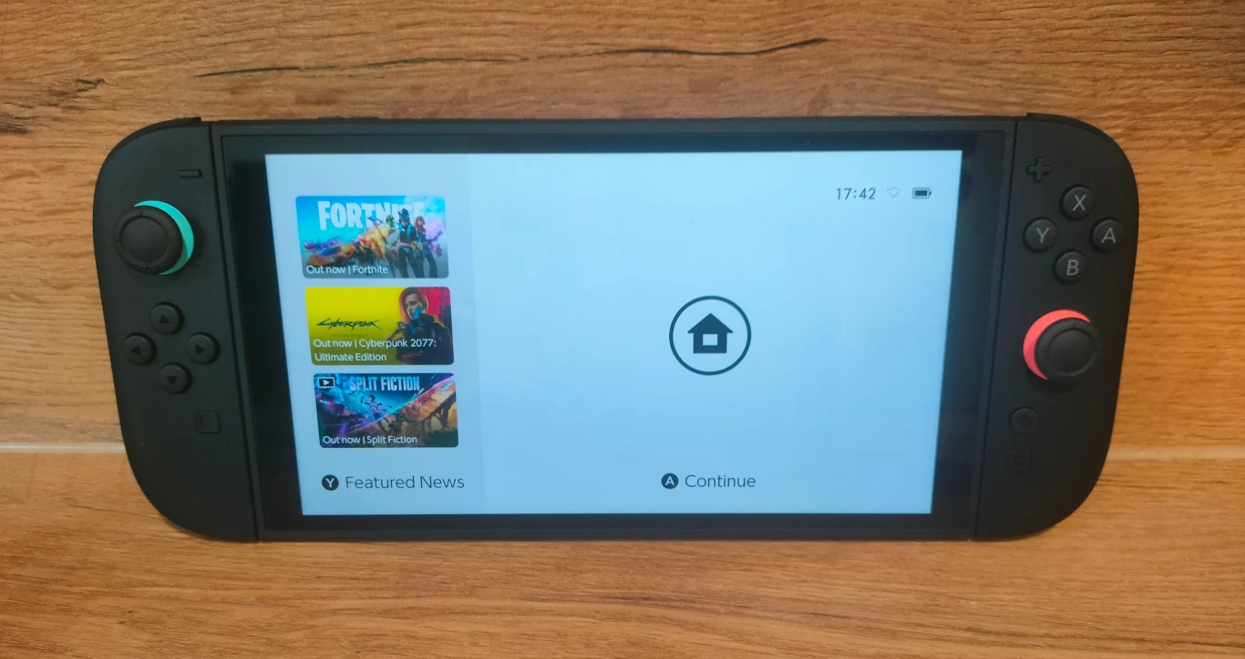 Konsola do gier Nintendo Switch leżąca na drewnianej powierzchni, ekran urządzenia wyświetla menu z aktualnościami oraz ikoną przejścia do ekranu głównego, po lewej stronie widoczne kolorowe przyciski joysticka.