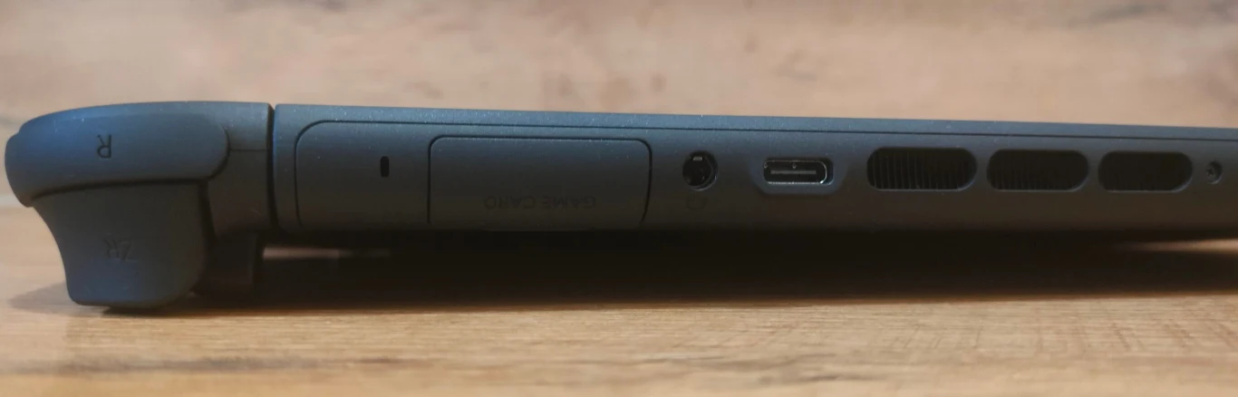 Boczny widok konsoli przenośnej z widocznym wejściem USB-C, kratkami wentylacyjnymi, wejściem słuchawkowym oraz fragmentem kontrolera po lewej stronie na tle drewnianej powierzchni.