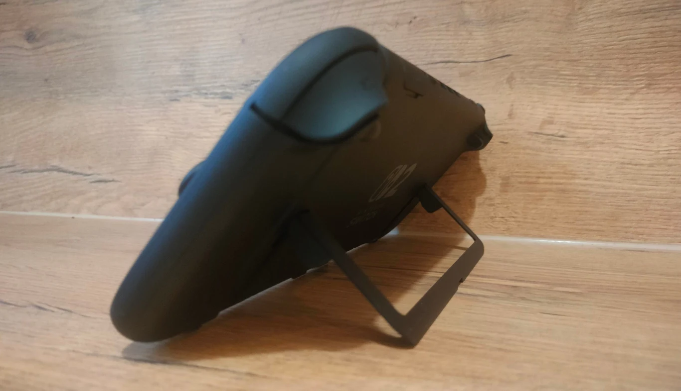 Konsola przenośna oparta na podpórce, ustawiona na drewnianej powierzchni, widoczna z boku, z wysuniętą nóżką służącą jako podstawa.