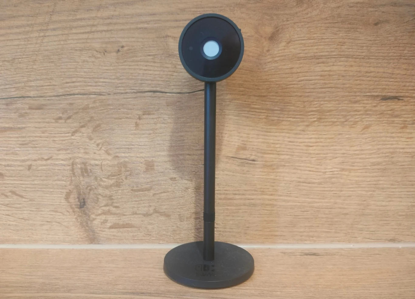 Czarna kamera internetowa na smukłej podstawie ustawiona na tle drewnianej ściany, konstrukcja minimalistyczna, nowoczesny design.