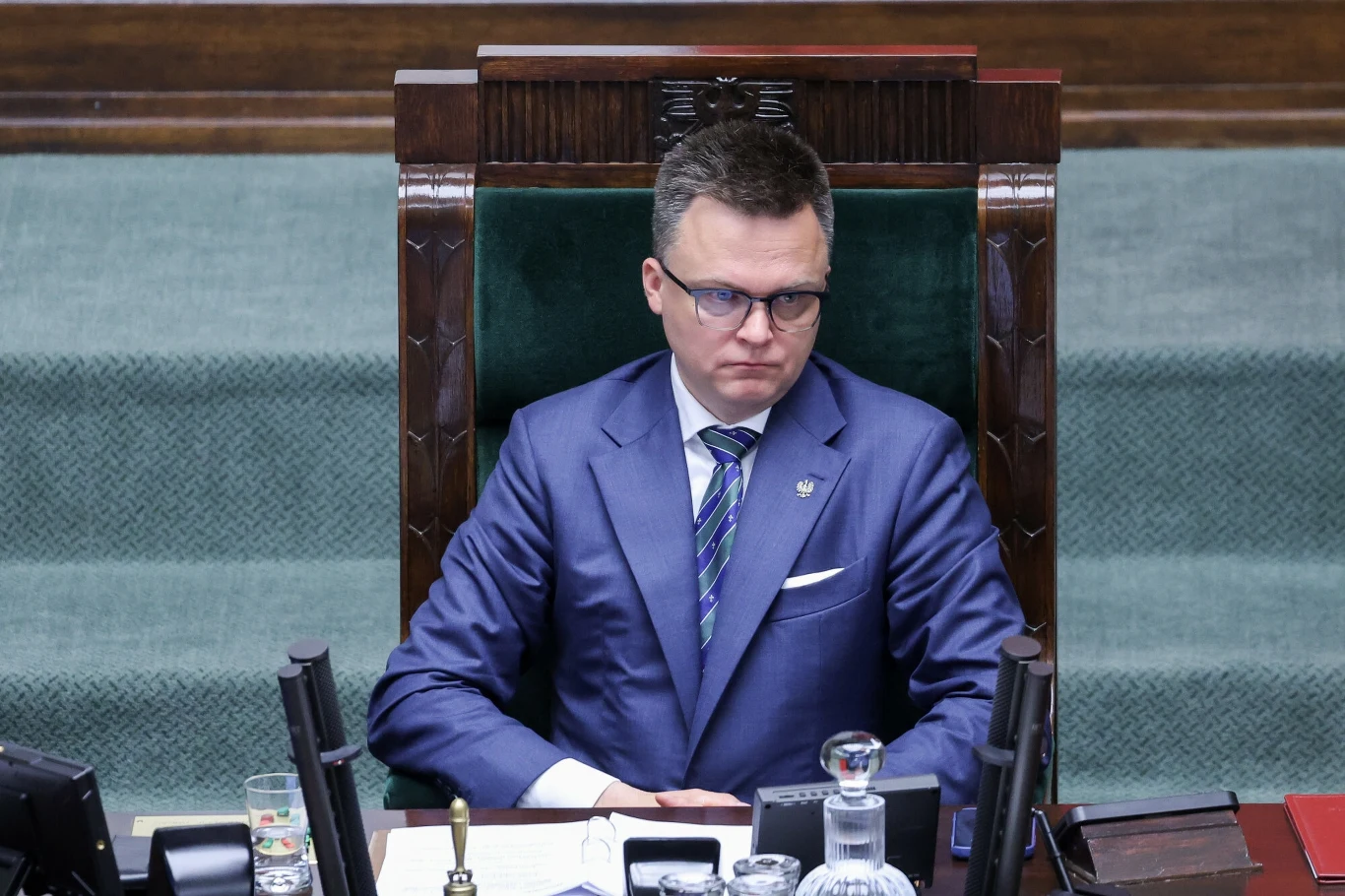 Marszałek Sejmu Szymon Hołownia Mężczyzna w garniturze siedzi w fotelu przewodniczącego w sali obrad, przed nim znajdują się mikrofony, dokumenty i szklana karafka.