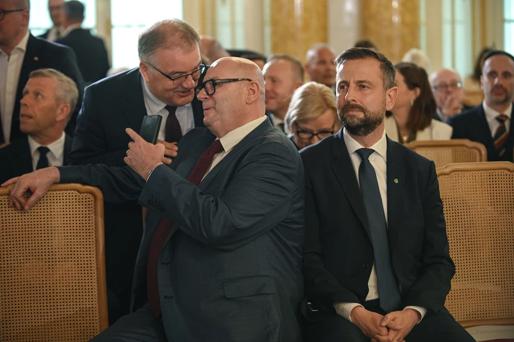 Politycy PSL Piotr Zgorzelski i Władysław Kosiniak-Kamysz na ceremonii wręczenia uchwały PKW Karolowi Nawrockiemu Grupa elegancko ubranych mężczyzn siedząca w rzędzie podczas oficjalnego wydarzenia, dwóch z nich prowadzi rozmowę i gestykuluje przy pomocy smartfona, reszta uczestników zwrócona jest w stronę sceny, panuje poważna atmosfera