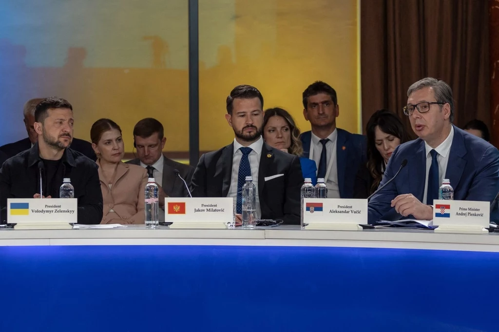 Wołodymyr Zełenski, prezydent Czarnogóry i Aleksandar Vuczić na szczycie w Odessie Trzech mężczyzn siedzi przy stole konferencyjnym, przed każdym z nich stoi karta z flagą oraz imieniem i nazwiskiem, co sugeruje oficjalne spotkanie międzynarodowe. Pozostali uczestnicy znajdują się w tle, skupiając uwagę na wypowiedzi jednego z lideró...