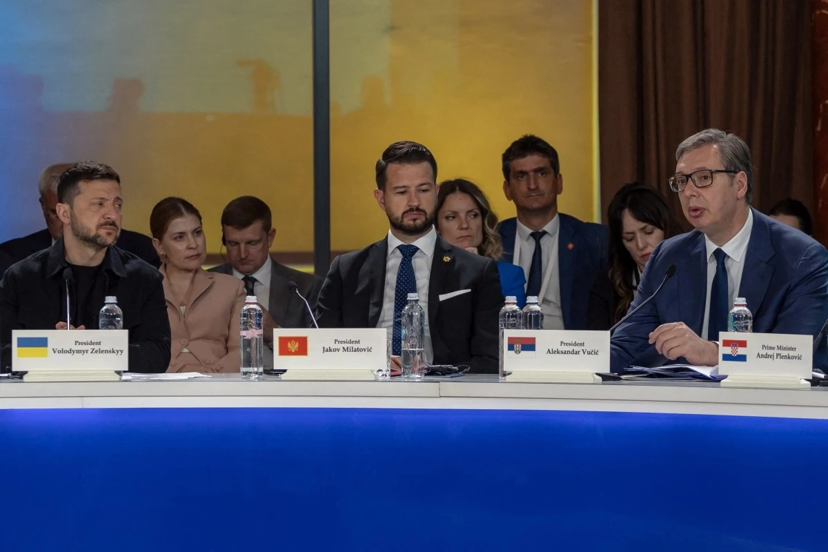 Trzech mężczyzn siedzi przy stole konferencyjnym, przed każdym z nich stoi karta z flagą oraz imieniem i nazwiskiem, co sugeruje oficjalne spotkanie międzynarodowe. Pozostali uczestnicy znajdują się w tle, skupiając uwagę na wypowiedzi jednego z lideró...