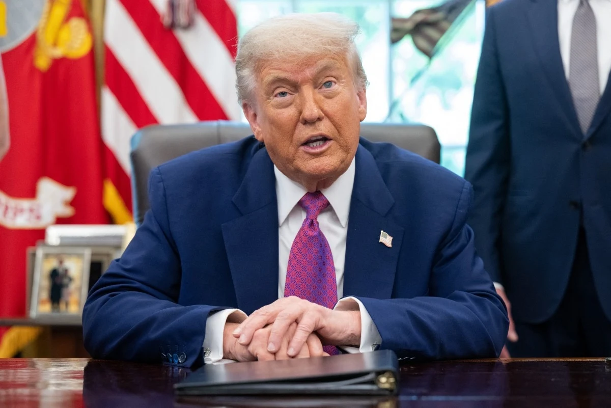 Prezydent USA Donald Trump Mężczyzna w niebieskim garniturze z różowym krawatem siedzi przy biurku w gabinecie, w tle widoczne są flagi oraz fragmenty wyposażenia wnętrza.