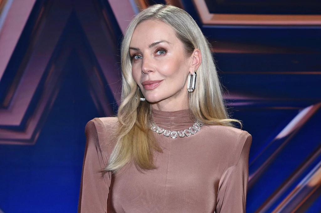 Kobieta z długimi blond włosami ubrana w elegancką, satynową suknię w jasnym kolorze, ozdobiona dużymi, efektownymi kolczykami i naszyjnikiem, stoi na tle ciemnoniebieskiego, geometrycznego motywu.
