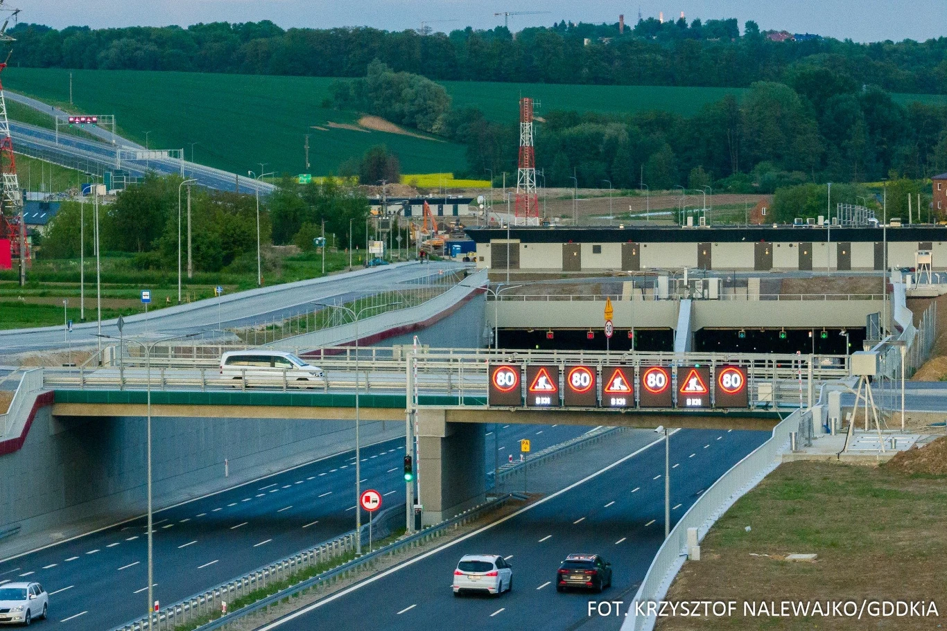 Wielopoziomowe skrzyżowanie autostrad z mostem, tunelem oraz elektronicznymi oznaczeniami ograniczenia prędkości nad jezdnią, wokół zielone pola i infrastruktura drogowa.