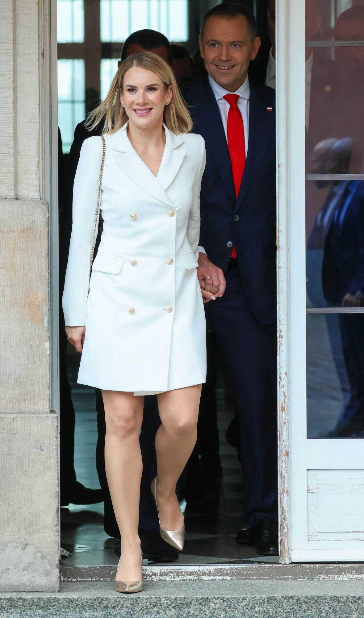 Marta Nawrocka przyciągnęła uwagę swoją stylizacją. Miała ją na sobie już drugi raz Kobieta w eleganckiej, białej marynarce sukienkowej wychodzi z budynku, trzymając za rękę mężczyznę w garniturze z czerwoną krawatą; oboje uśmiechnięci, w tle fragment drzwi i okno.