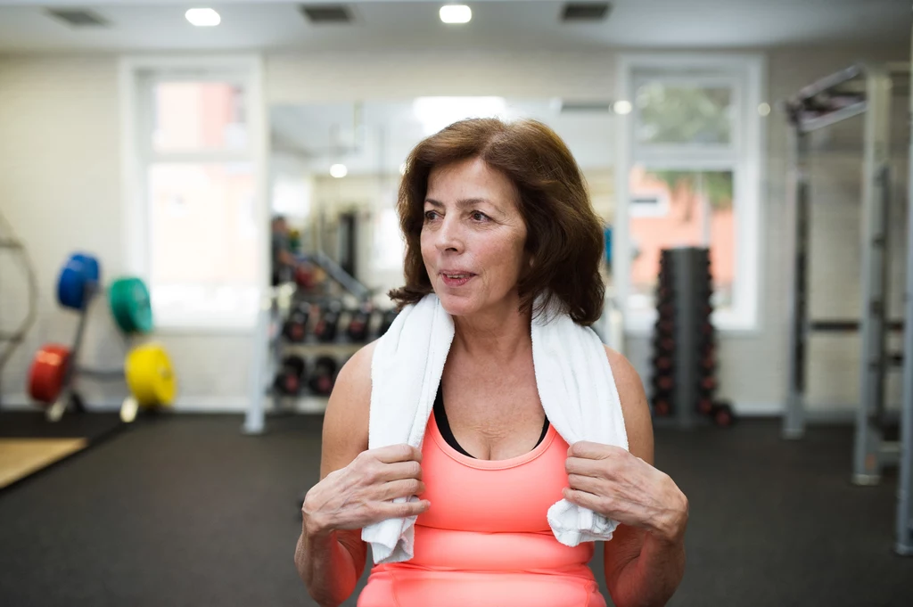 Badania naukowe potwierdzają, że kobiety po menopauzie zyskują ogromne korzyści ze stosowania treningu siłowego Kobieta w średnim wieku w sportowym stroju stoi na siłowni z ręcznikiem przerzuconym przez ramiona, w tle widoczne hantle oraz sprzęt do ćwiczeń.