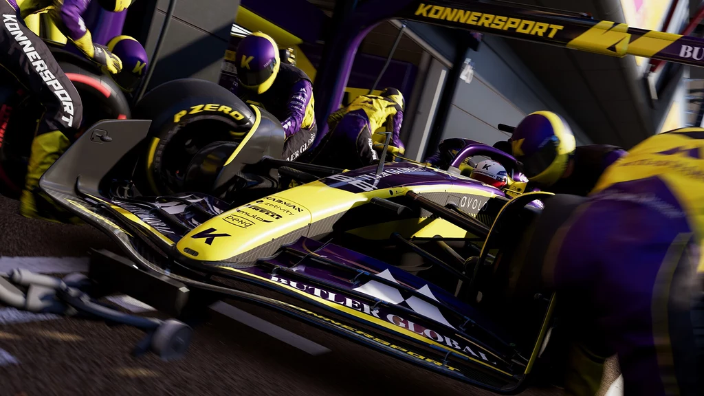 F1 25 Zespół mechaników podczas pit stopu obsługuje samochód wyścigowy Formuły 1 w żółto-fioletowych barwach, w tle widoczny garaż zespołu oraz fragment toru.