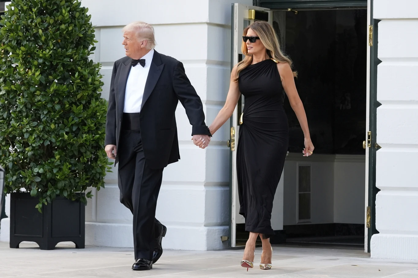 Elegancko ubrana para trzymająca się za ręce wychodzi z budynku o jasnej fasadzie, kobieta w czarnej sukni i okularach przeciwsłonecznych, mężczyzna w klasycznym smokingu.