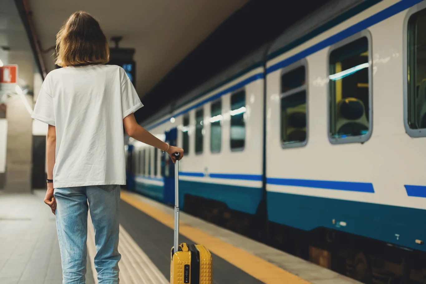 W przedziale nie działa klimatyzacja? Masz prawo do wniesienia reklamacji Osoba z jasnymi włosami ubrana w biały T-shirt i niebieskie jeansy stoi na peronie z żółtą walizką na kółkach, patrząc w stronę stojącego obok pociągu.