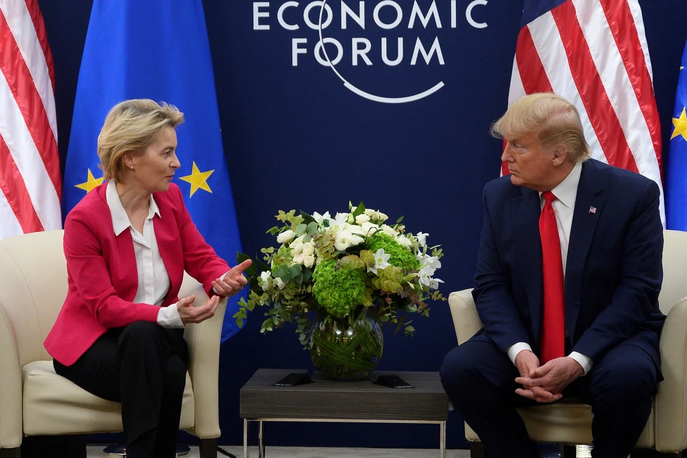 Szefowa KE Ursula von der Leyen i prezydent USA Ursula von der Leyen Dwie osoby siedzące naprzeciwko siebie w eleganckich fotelach podczas rozmowy, po środku kompozycja kwiatowa na stoliku, w tle flagi Unii Europejskiej i Stanów Zjednoczonych oraz logo forum ekonomicznego.