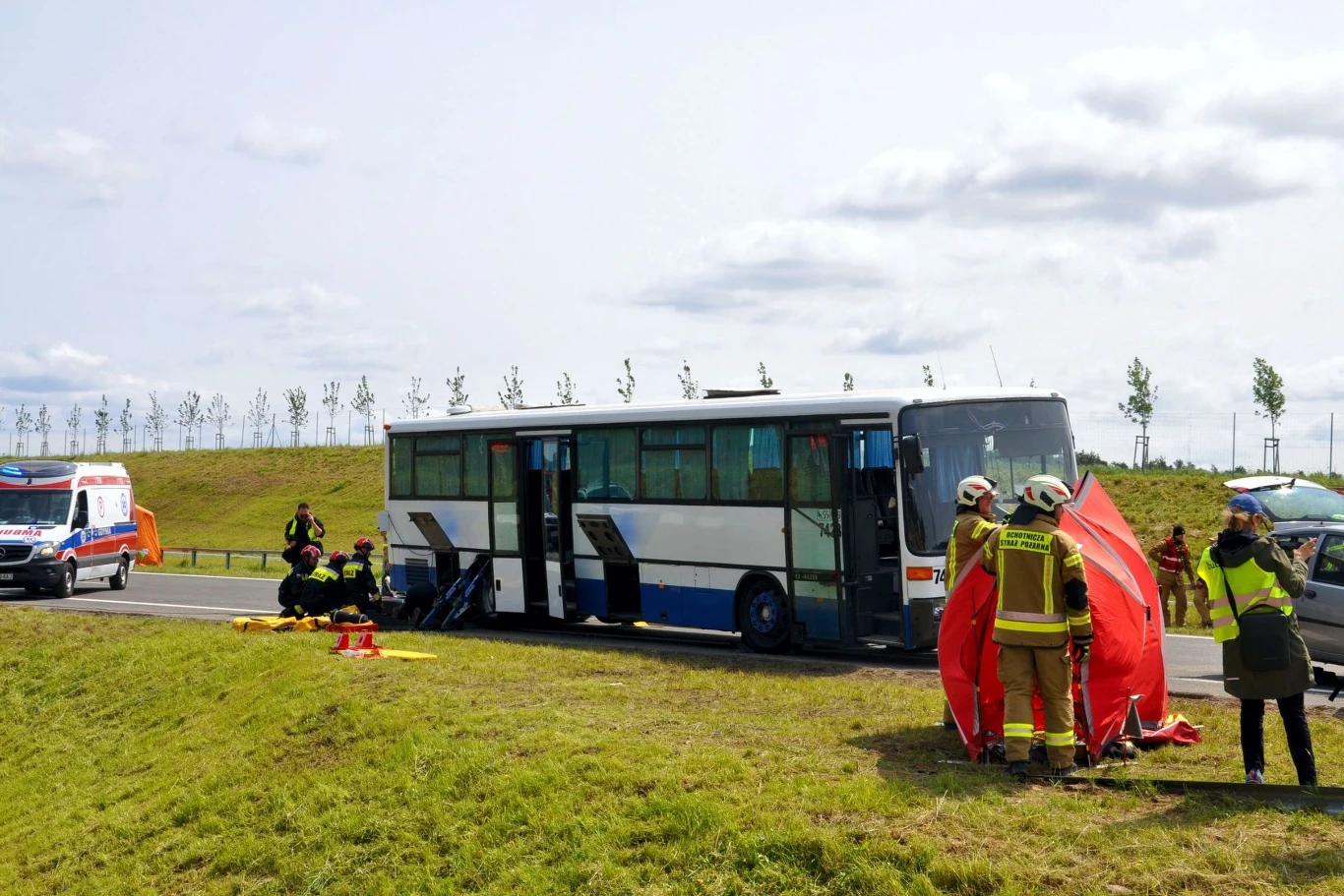 Akcja służb ratowniczych na Obwodnicy Metropolii Trójmiejskiej Służby ratownicze pracujące przy autobusie stojącym na poboczu drogi, strażacy rozstawiają czerwoną zasłonę oraz sprzęt medyczny, w tle widoczne karetki pogotowia.