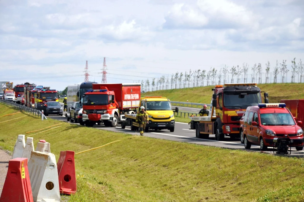 Służby w akcji na OMT. Odcinek drogi szybkiego ruchu przetestowany Kolumna pojazdów służbowych, w tym straży pożarnej, pomocy drogowej i policji, na autostradzie przy zamkniętym pasie ruchu, z ustawionymi barierkami bezpieczeństwa i zielonym pasem pobocza.