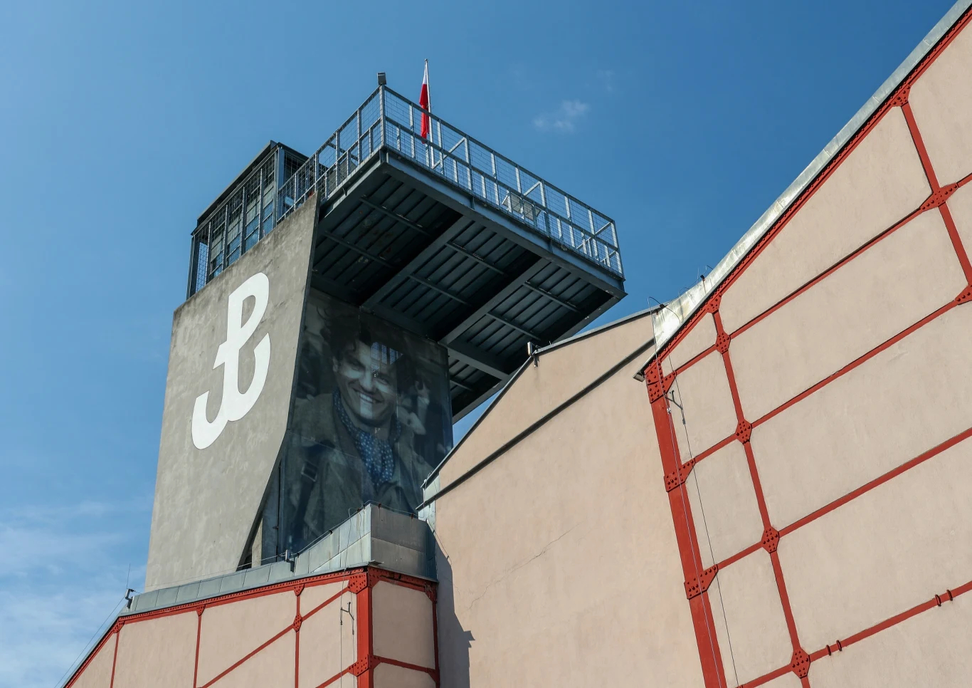 Muzeum Powstania Warszawskiego w czerwcu ogłosi przetarg na rozbudowę Fragment nowoczesnego budynku przemysłowego z widoczną dużą literą 'P' stylizowaną na kotwicę na szarej ścianie oraz muralem z twarzą, na dachu platforma widokowa z balustradą i polska flaga na tle nieba.