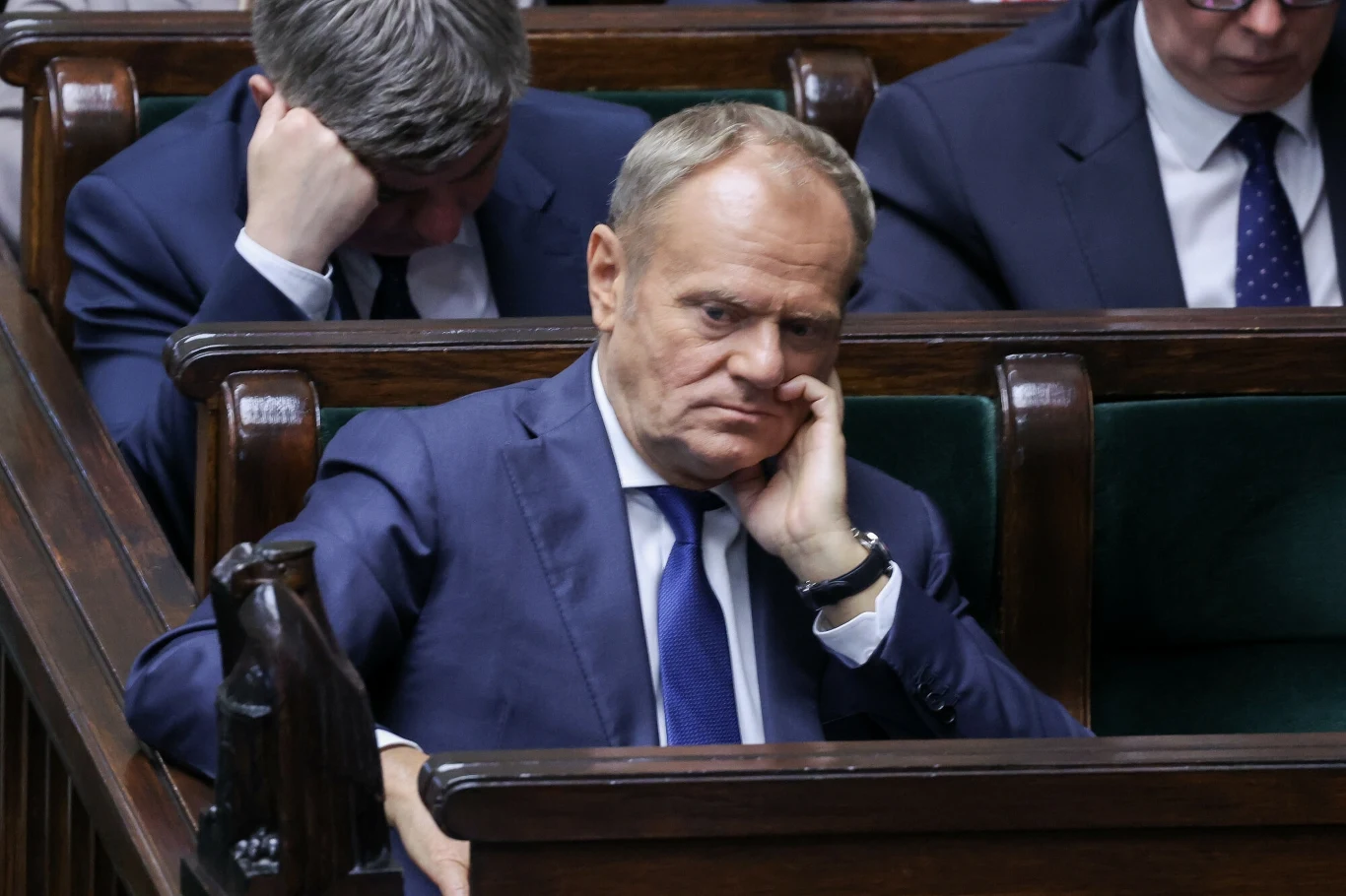 Premier Donald Tusk Mężczyzna w garniturze siedzący na ławie w parlamencie, oparty o rękę, z zamyślonym wyrazem twarzy, za nim inni mężczyźni w formalnych ubraniach również przyjmują zamyślone lub znudzone pozy.