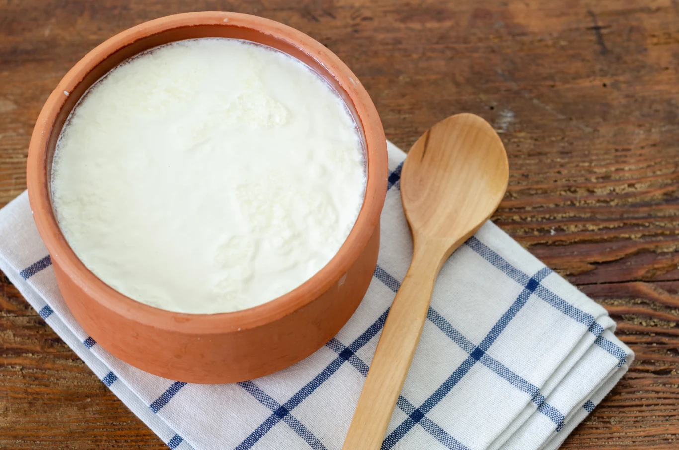 Gliniany garnuszek wypełniony jogurtem naturalnym ustawiony na złożonej, biało-niebieskiej ściereczce w kratę, obok drewniana łyżka, tło stanowi drewniany stół.