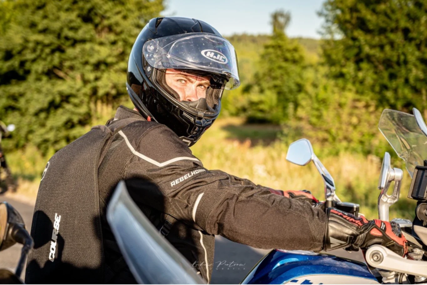 Przyjechałeś motocyklem lub skuterem bez kasku? Nie zatankujesz. Mężczyzna w czarnym kasku motocyklowym oraz kurtce motocyklowej siedzi na niebieskim motocyklu, patrząc w stronę aparatu, w tle widoczne drzewa i roślinność.