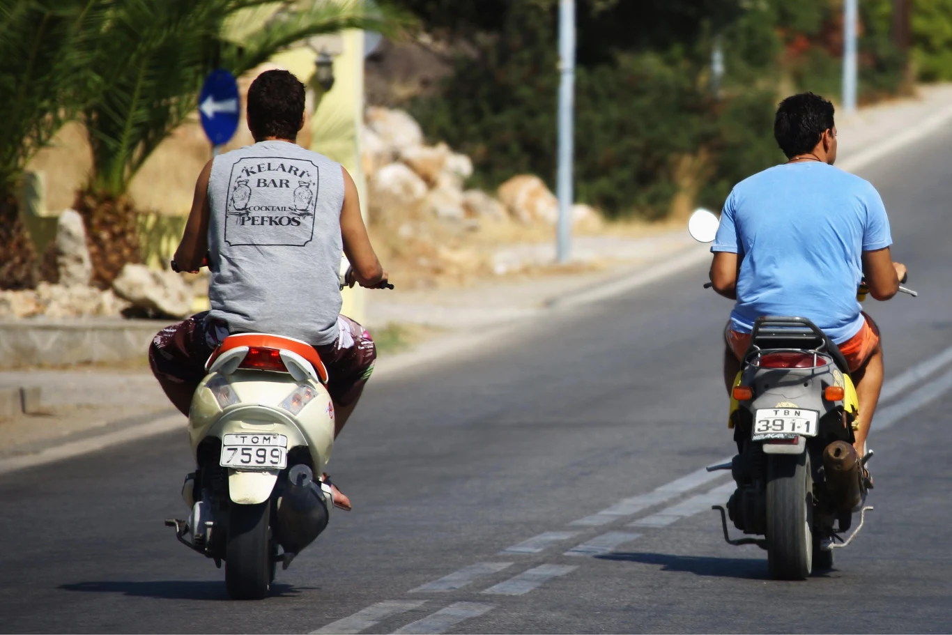 Jazda skuterem bez kasku w Grecji to zupełnie normalny widok. Nowe przepisy wejdą w życie we wrześniu bieżącego roku. Dwóch mężczyzn jadących na skuterach po pustej asfaltowej drodze w słoneczny dzień, jeden ubrany w szarą koszulkę bez rękawów, drugi w niebieską koszulkę z krótkimi rękawami, wokół roślinność i kamienne elementy na poboczu.