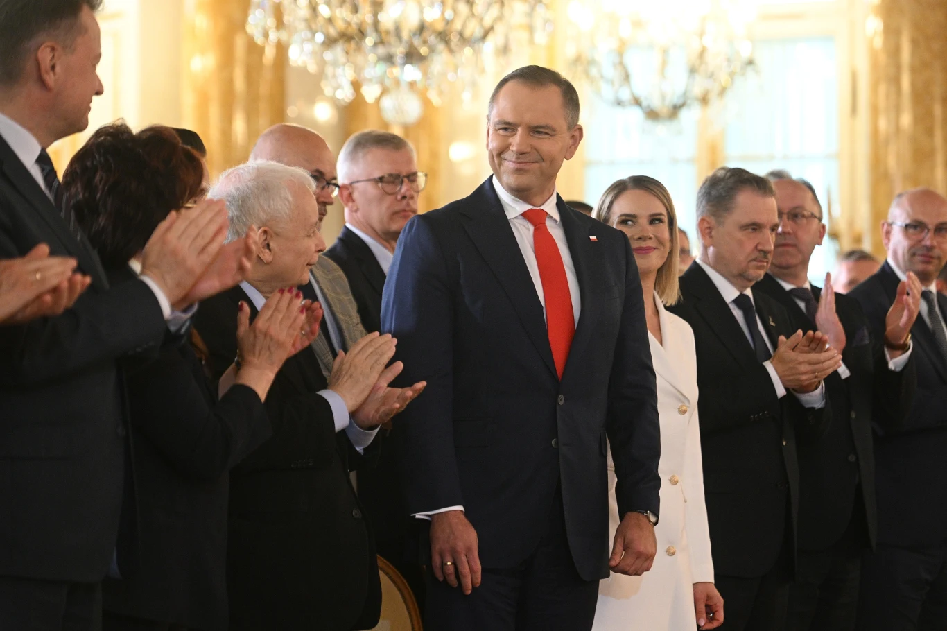 Karol Nawrocki, prezydent elekt podczas uroczystości przekazania przez PKW zaświadczenia o wyborze na prezydenta RP Grupa elegancko ubranych osób w jasnej, reprezentacyjnej sali, główną postacią jest mężczyzna w granatowym garniturze i czerwonym krawacie, wokół którego zgromadzeni ludzie biją brawo i patrzą na niego z uznaniem.